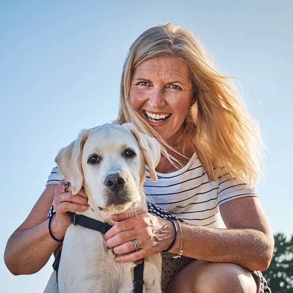Frau kniet lachend neben ihrem hellen Labrador im Gegenlicht
