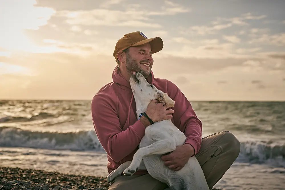 Mann umarmt seinen hellen Labrador am Ostseestrand bei Sonnenuntergang
